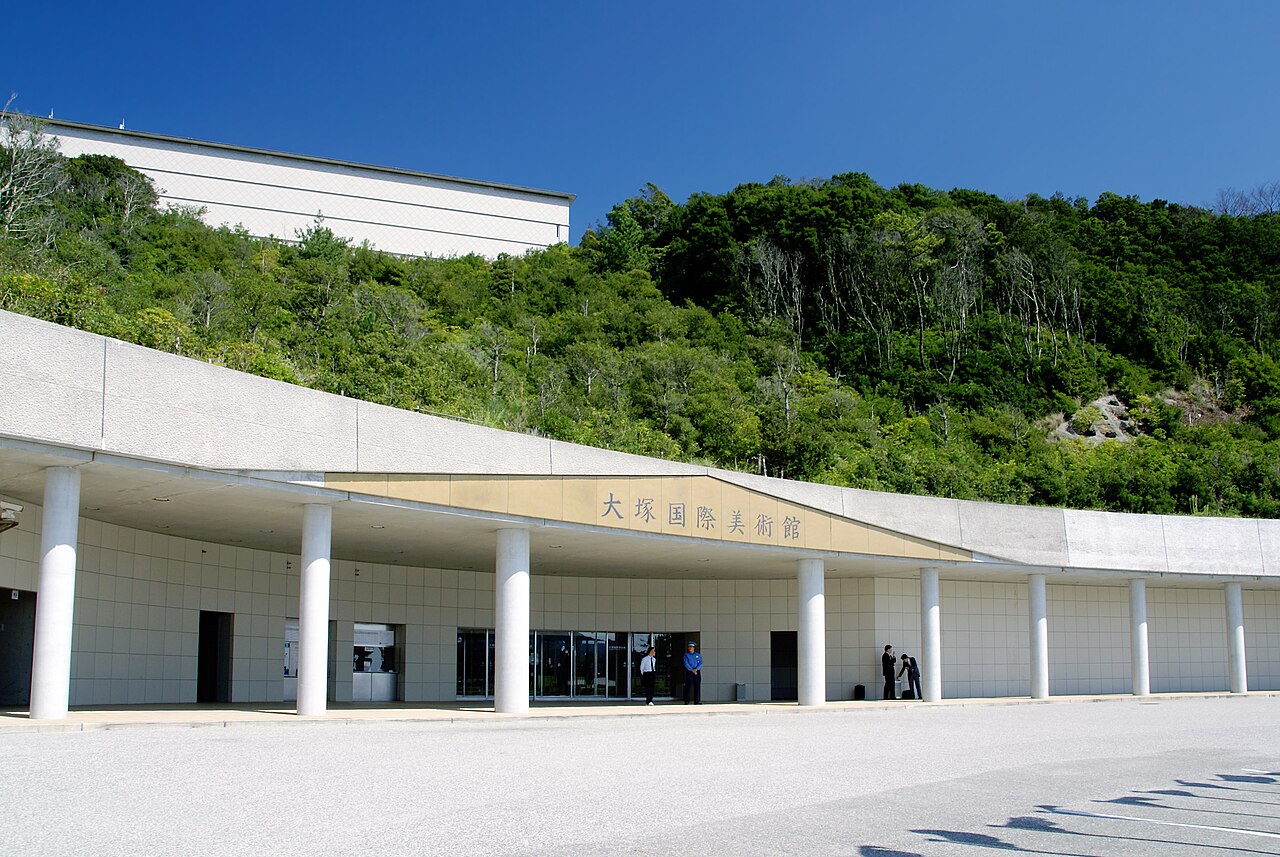 The low white colonnaded entrance of the Otsuka Museum of Art, with its name sign above the doors and a wooded hillside rising behind it.