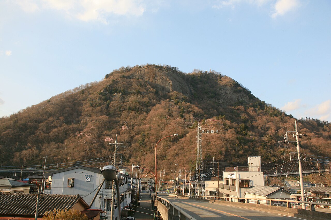 A steep rocky-faced mountain rises above a town with low houses, power pylons and a road bridge in the foreground under a pale blue sky.