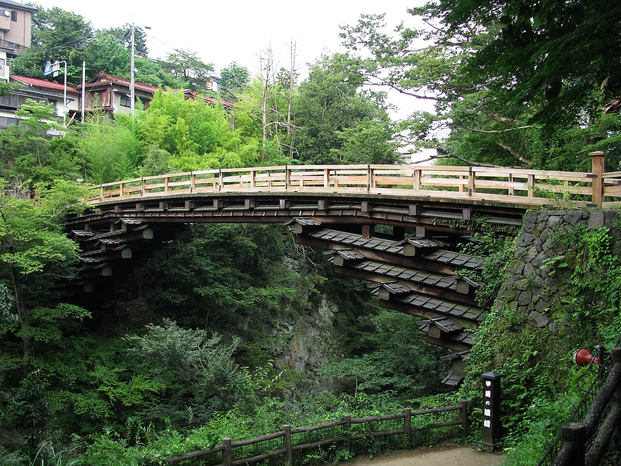 A wooden arched bridge with a railed deck and rows of tiled-roof cantilever beams spans a green wooded gorge, with houses on the slope behind and a small sign post at the lower right.