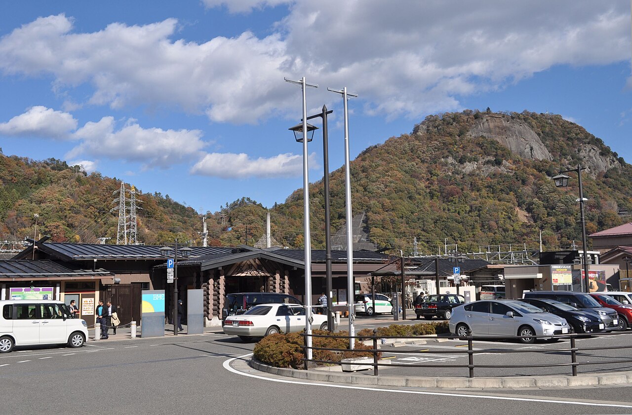 A station building with a timber-and-log entrance and a bus and taxi forecourt sits below wooded hills and a rocky-faced mountain under a blue sky with large white clouds.