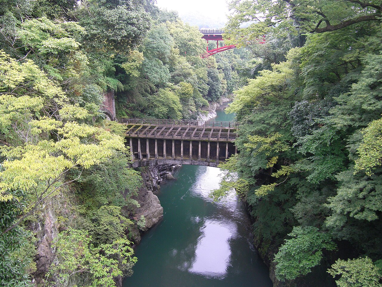 A concrete aqueduct bridge crosses a deep green river gorge between wooded cliffs, with a red steel bridge visible far upstream in the haze.