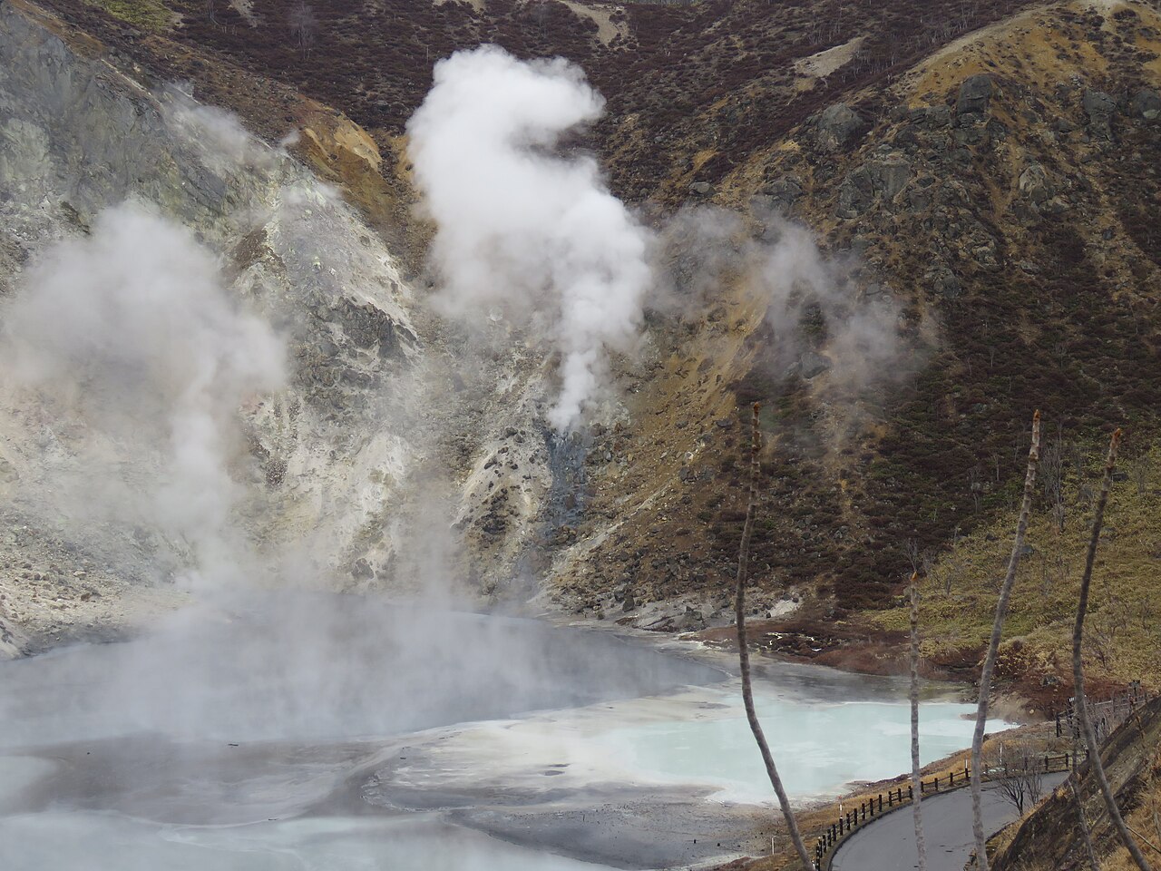 Oyunuma, a steaming sulphur pond below Noboribetsu Onsen, with columns of white vapour rising against a bare hillside and a path running along the far shore.