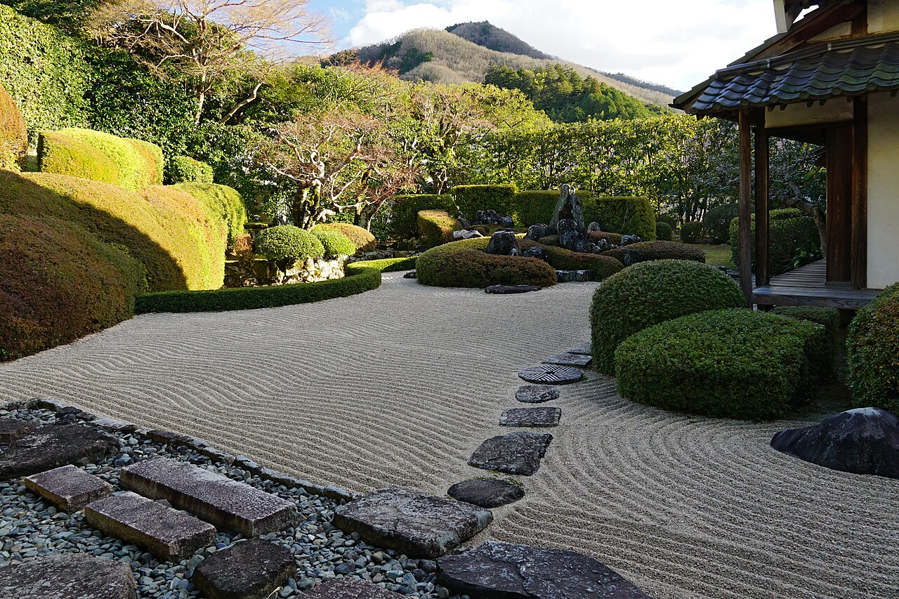 The dry landscape garden of Raikyuji Temple with raked gravel, stepping stones and clipped azalea mounds, with a mountain beyond the hedge