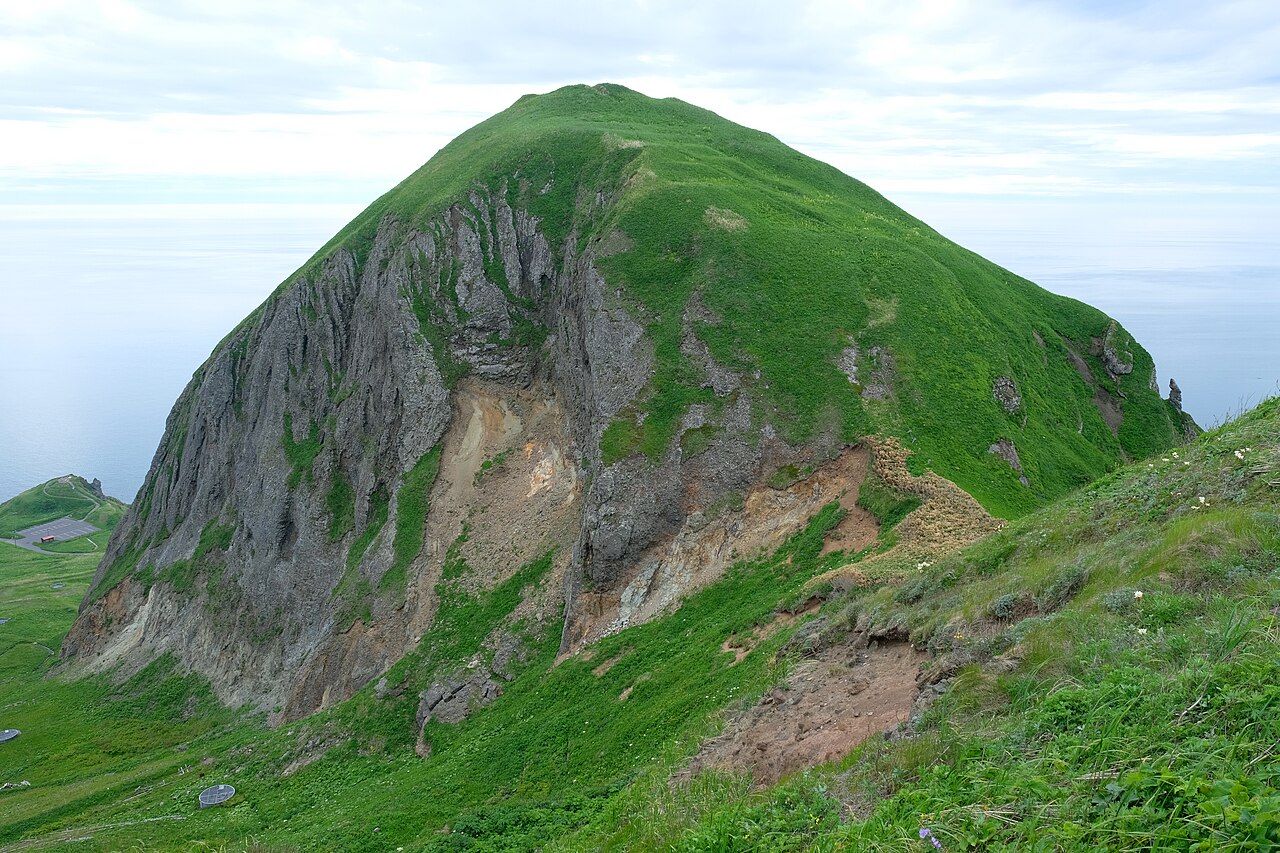 A steep grey rock dome covered in green grass on its upper slopes rises against a pale sky above Momoiwa on Rebun Island.