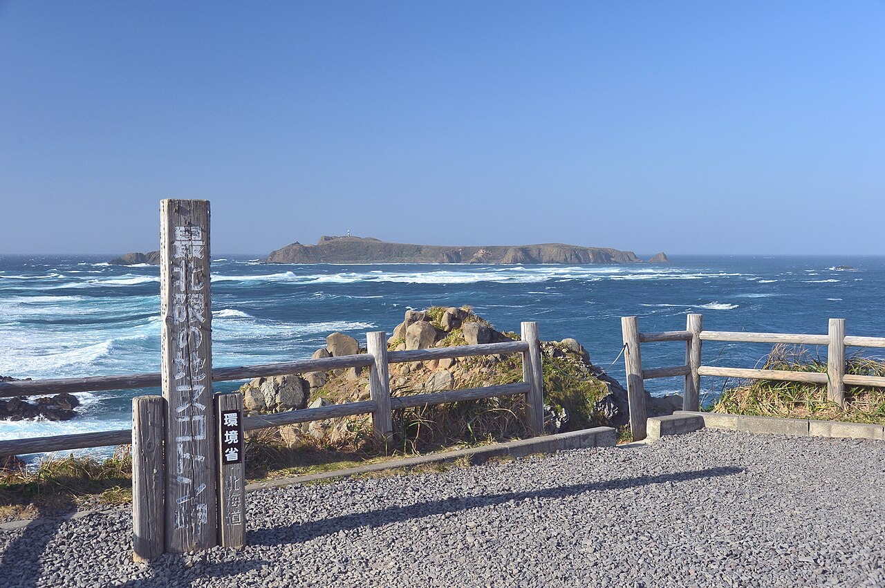A tall wooden monument post stands beside a fenced gravel viewpoint above rough blue sea with a low island on the horizon at Cape Sukoton.