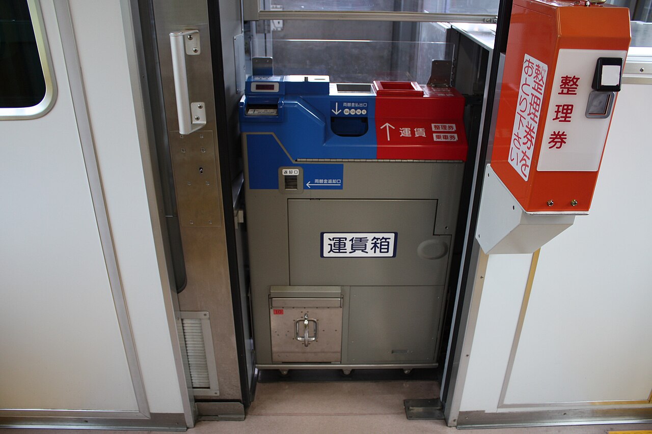 Inside a one-man train by the door: a blue-and-red fare box, with an orange numbered-ticket dispenser mounted on the wall beside it.