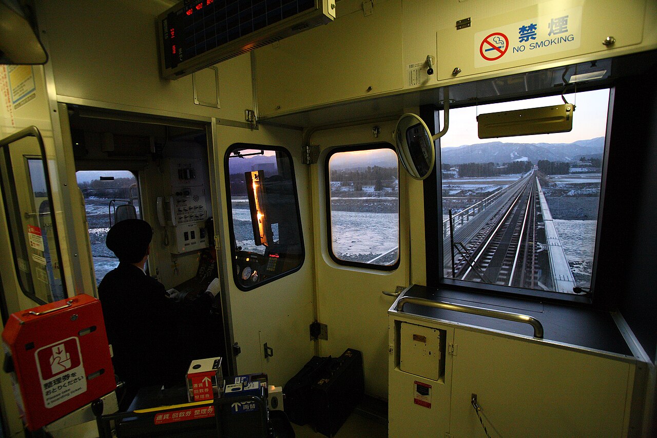 The front of a one-man train, with the driver at the controls, a fare box by the door and a snowy valley ahead through the windscreen.