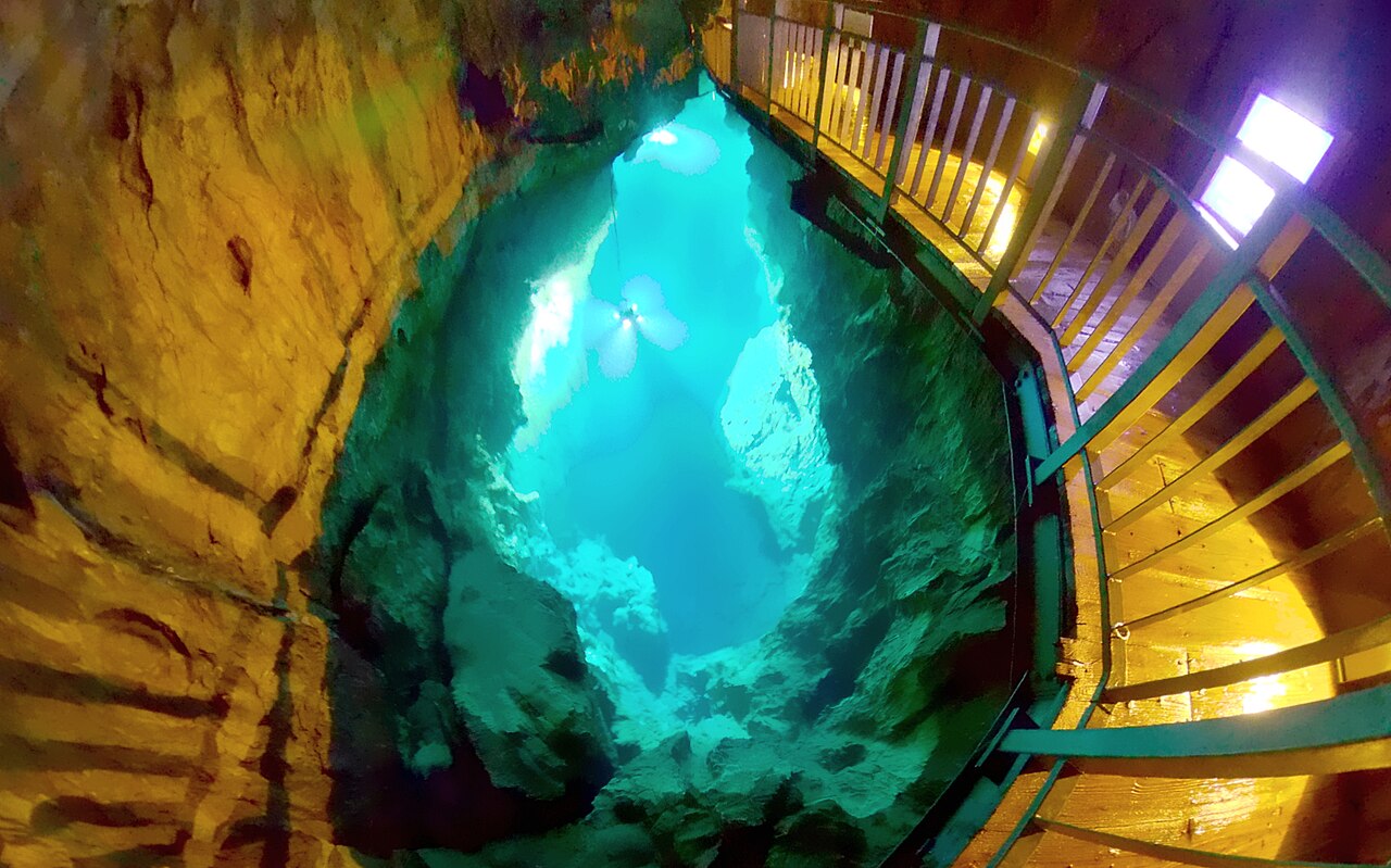 Turquoise underground lake seen from a wooden walkway inside Ryusendo Cave, with orange limestone walls around the water.