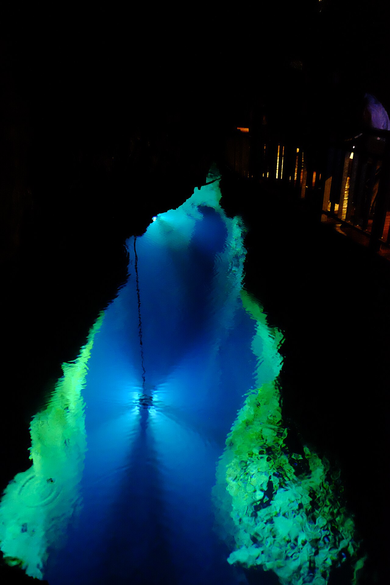 Deep blue underground lake in Ryusendo Cave, lit from below with green-lit rock edges and a wooden walkway at the right.