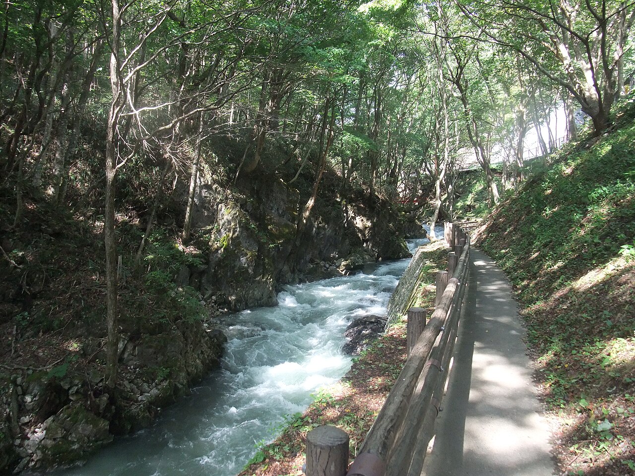 A fast clear stream running through a wooded gorge beside a paved footpath near Ryusendo Cave.