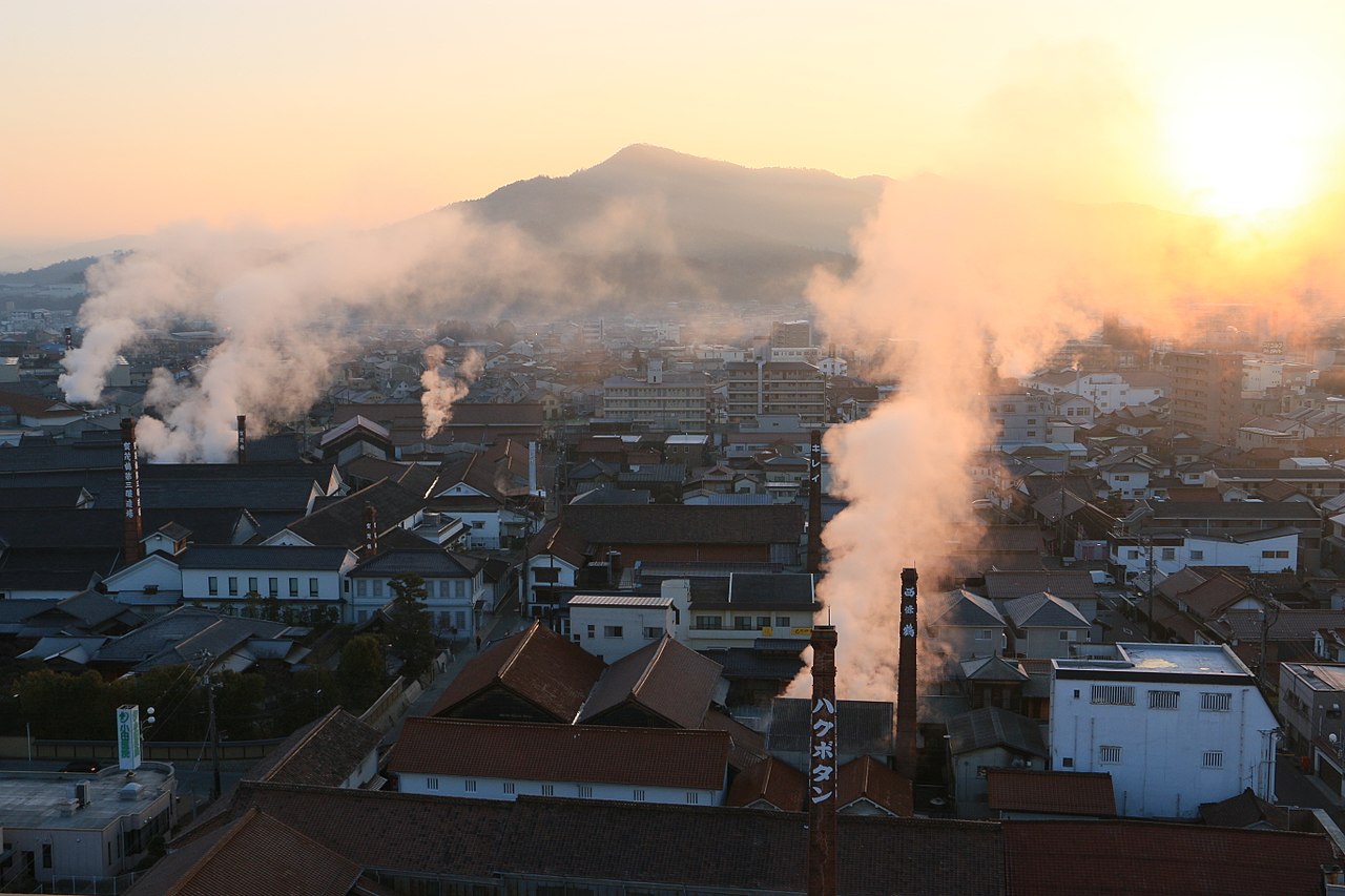 Sunrise over the Saijo brewery district, with several tall brick chimneys and plumes of steam rising above the rooftops and a mountain behind.