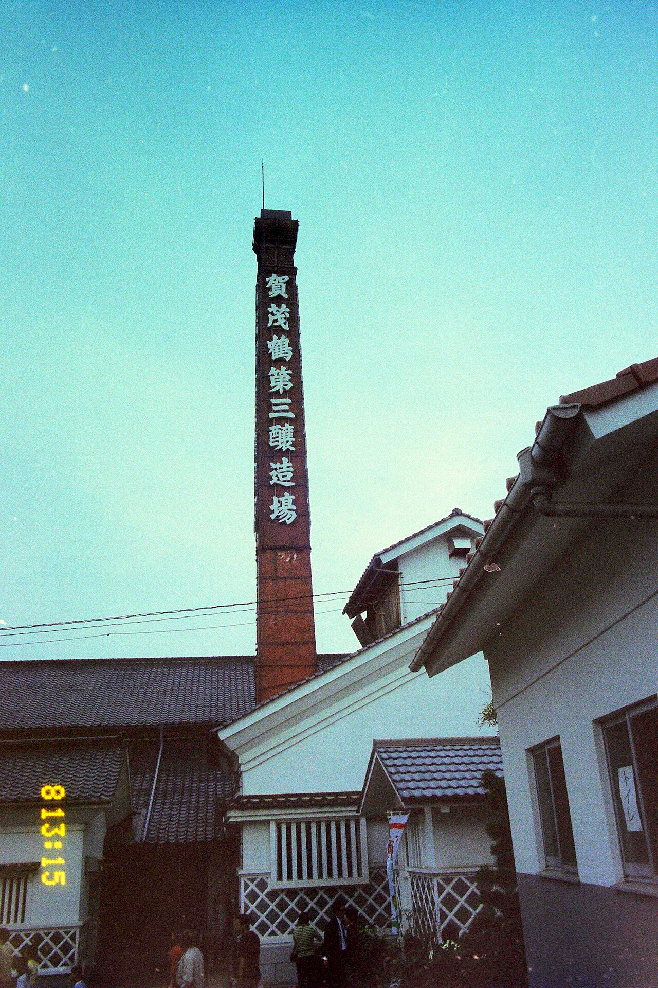 A tall brick brewery chimney with vertical lettering rising above the tiled roofs and white walls of a Saijo sake brewery under a clear sky.