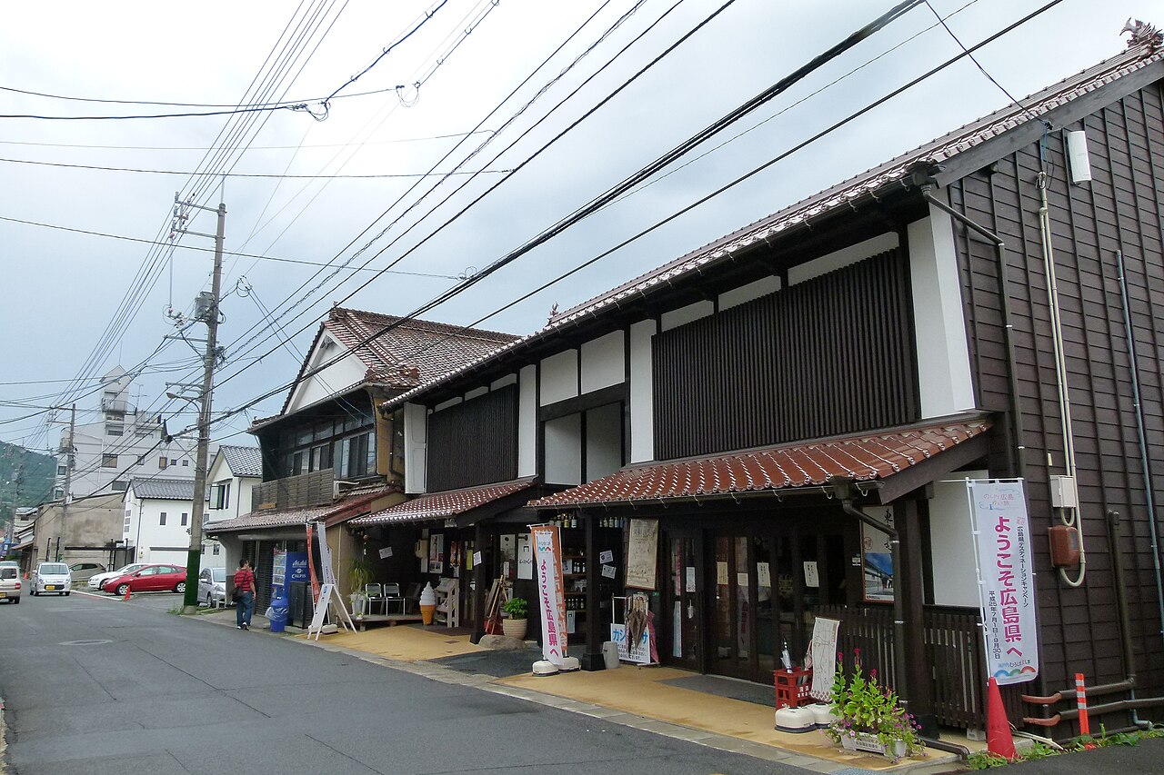 A traditional two-storey brewery building with a tiled roof and wooden facade on a Saijo street, with a shop entrance, banners and a car parked along the road.