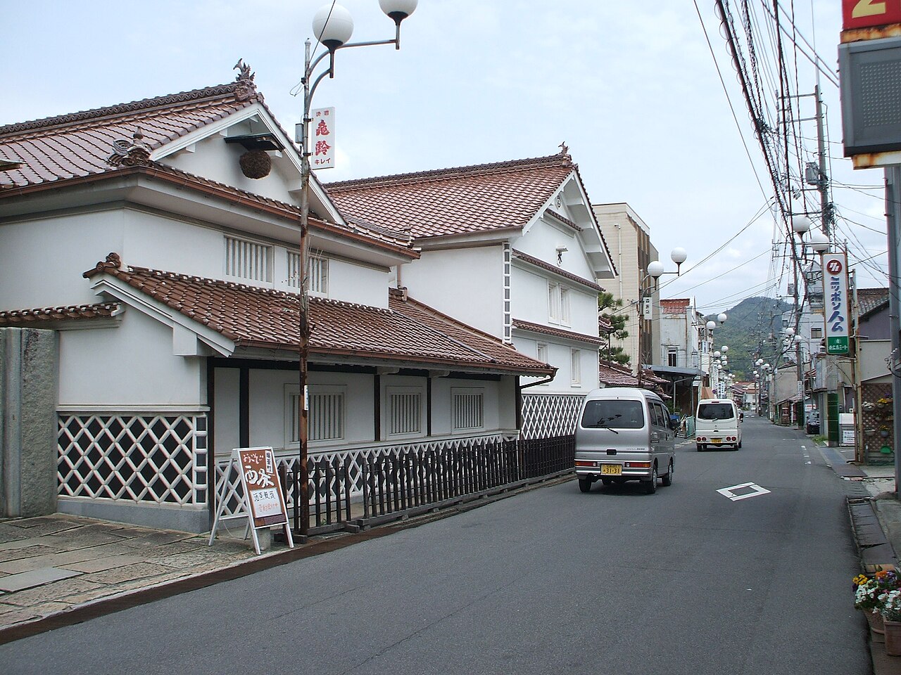 A street in the Saijo sake district, lined with white-walled brewery storehouses with red-tiled roofs and black lower walls, with utility poles overhead and hills in the distance.