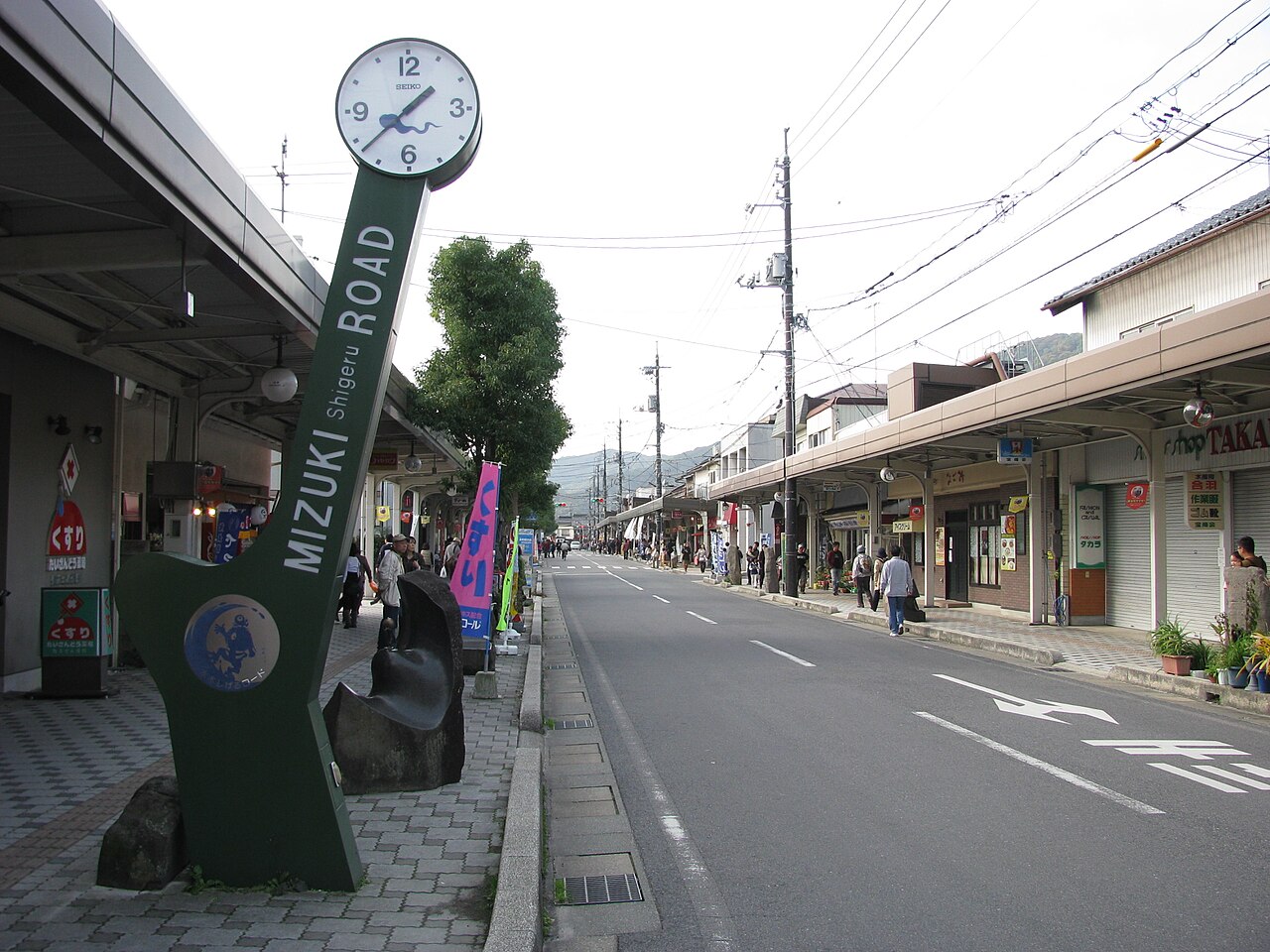 Green Mizuki Shigeru Road clock pillar with a yokai emblem beside a shopping street under covered arcades, with a few people walking toward distant hills
