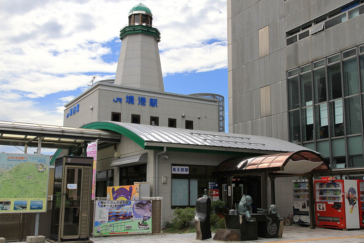JR Sakaiminato Station building topped with a small lighthouse-style tower, with a group of figures at the entrance under a partly cloudy sky
