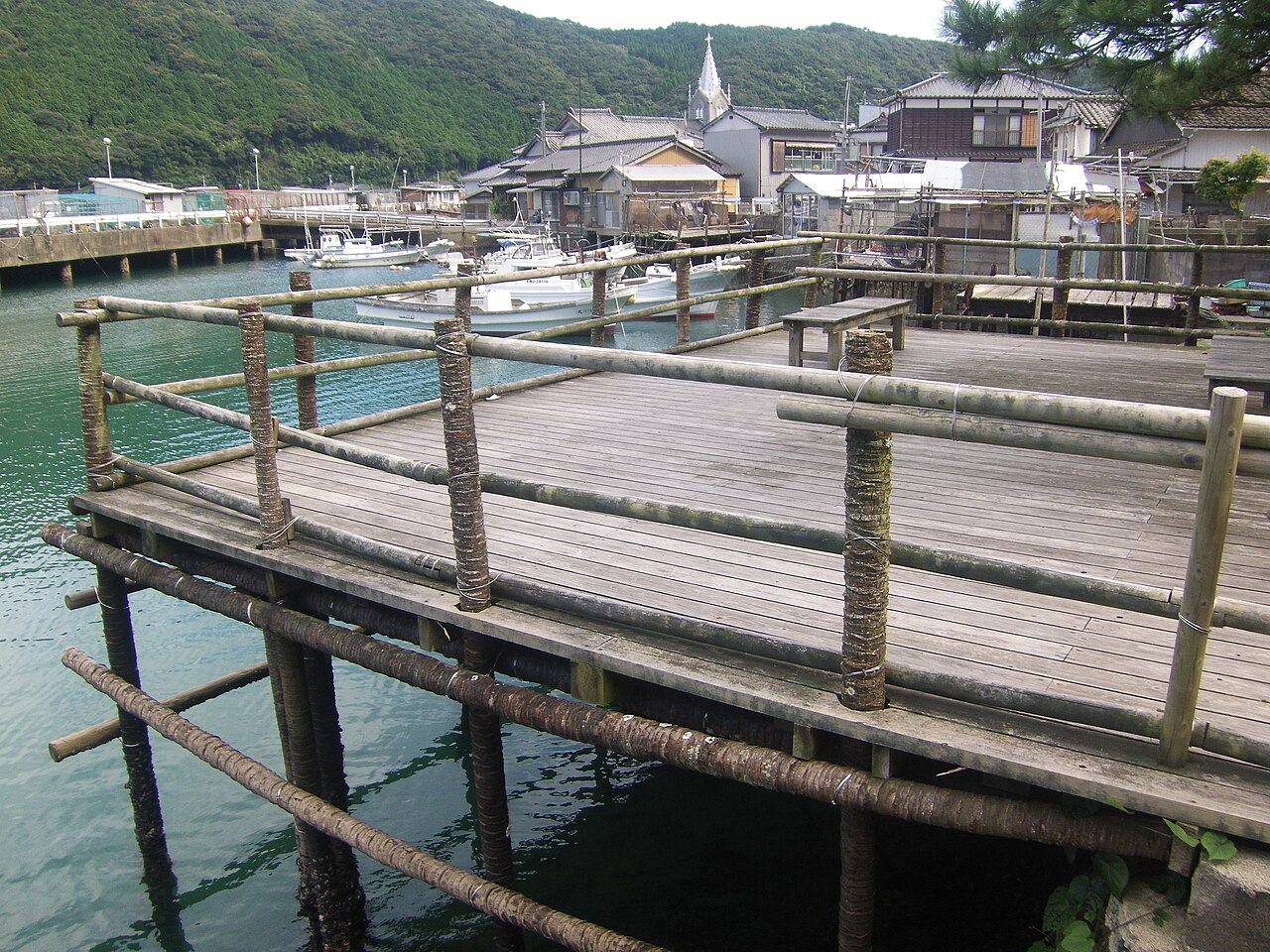 A wooden kake work platform on stilts over the water, with fishing boats and the Sakitsu church spire across the inlet.