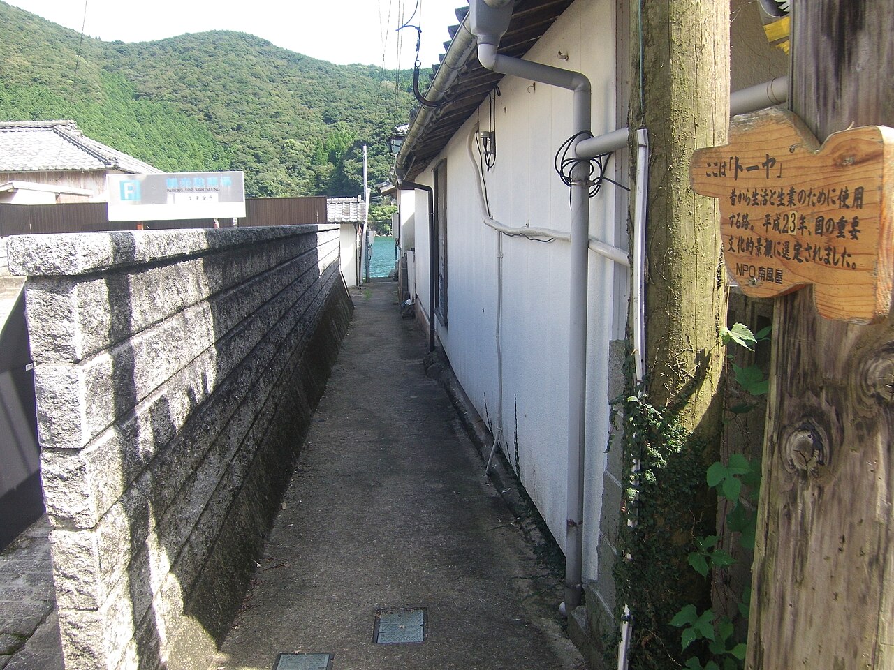 A narrow touya lane between houses in Sakitsu, with a wooden information sign and the water at the far end.