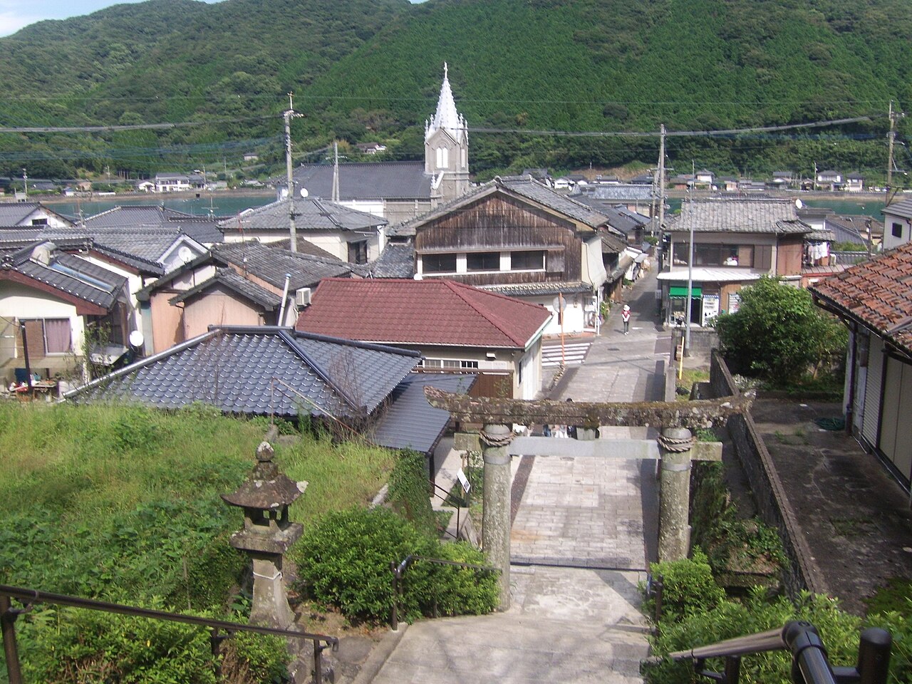 Tiled roofs of Sakitsu village below green hills, with a stone torii in the foreground and the church spire in the distance.