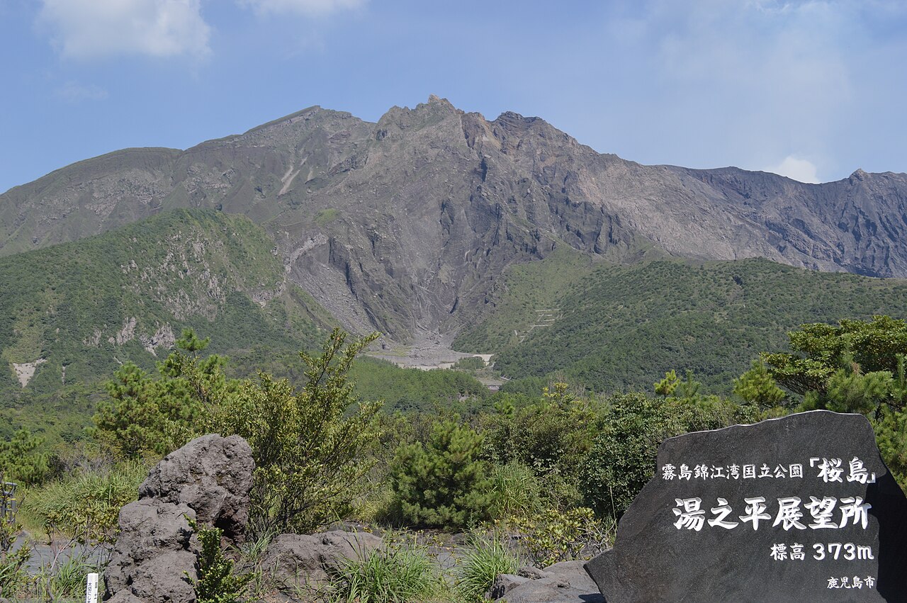 The jagged grey summit of Sakurajima above green scrub, with a carved stone marker for the Yunohira Observatory in the foreground