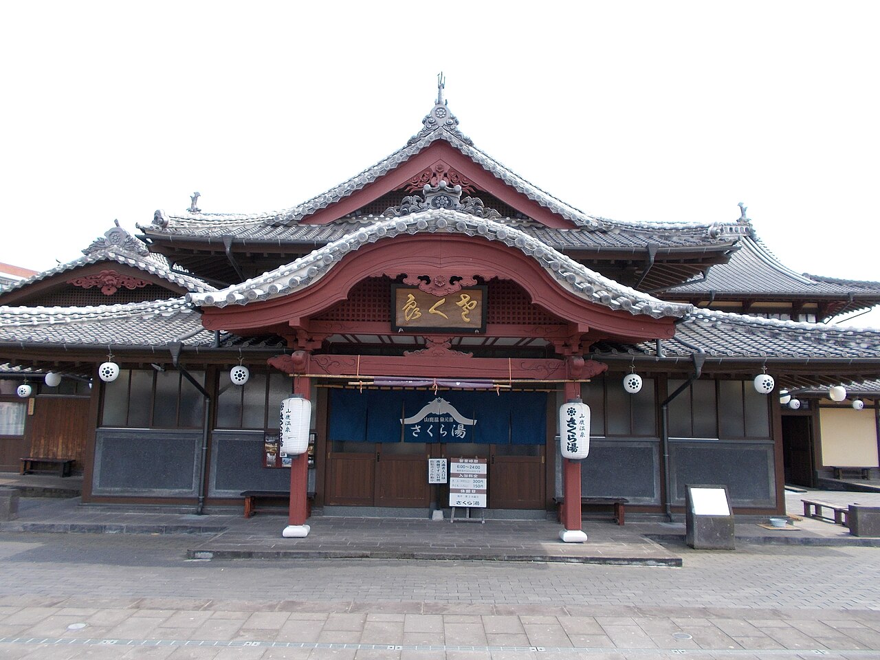 The red-lacquered curved gable of the Sakura-yu public bathhouse in Yamaga, with a dark indigo curtain across the entrance and white paper lanterns on either side.