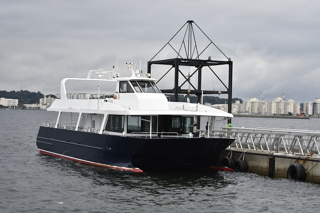 A dark-hulled passenger ferry moored at a pier with a crane frame behind it, the kind of boat used for the short crossing to Sarushima