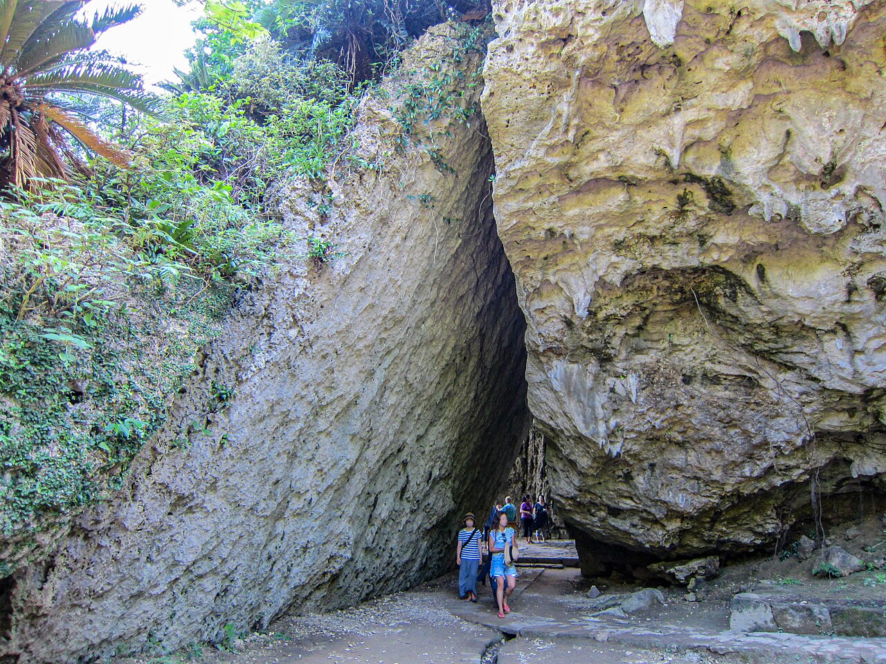Two huge slabs of rock leaning against each other to form a triangular passage at Sefa Utaki, with a few people walking beneath.