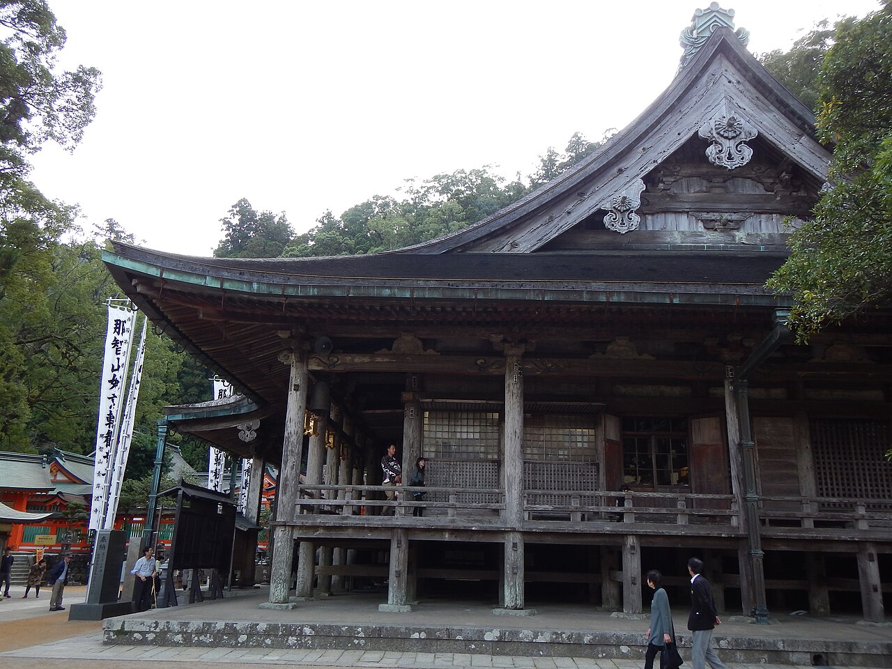 The weathered dark-wood main hall of Seiganto-ji, with a deep curved tiled roof and open veranda, stands among trees with a few visitors walking past below cloth banners.