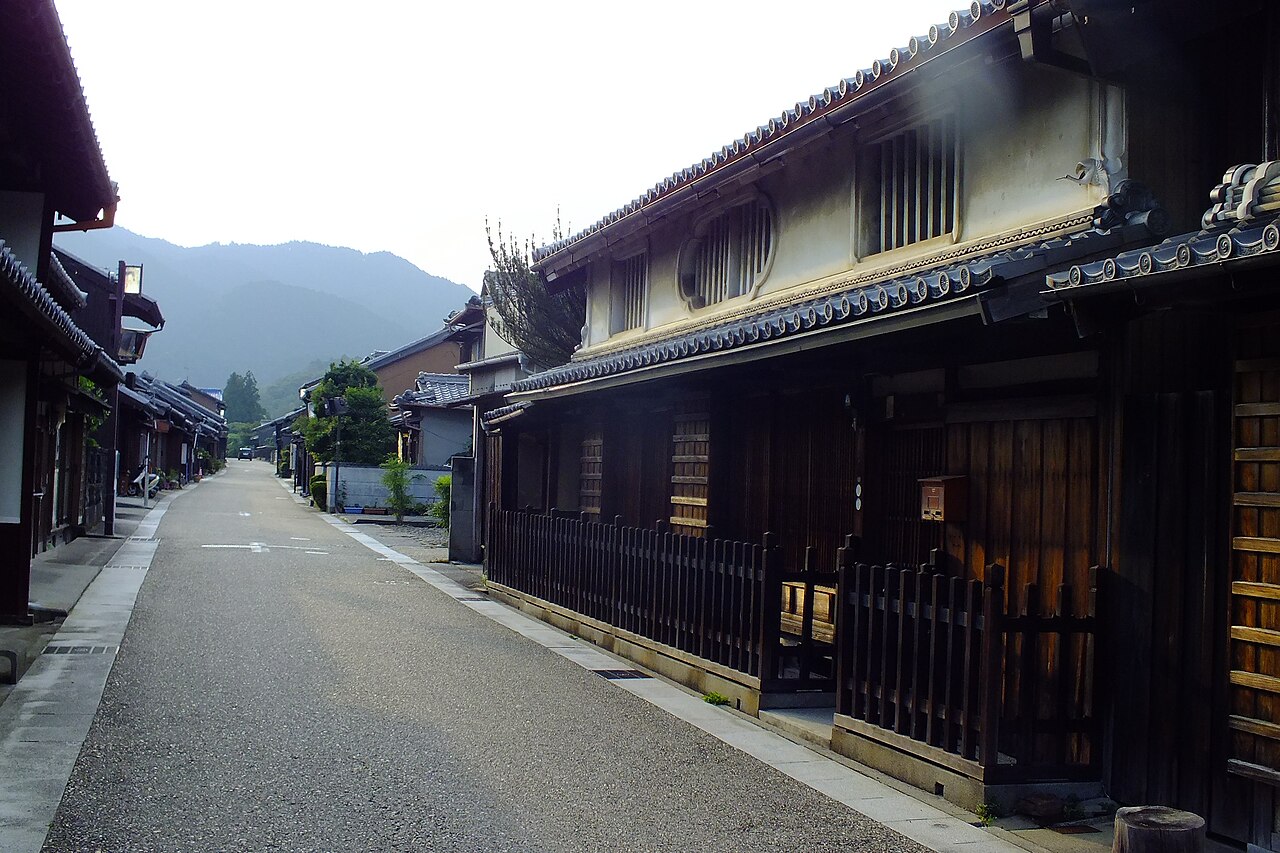 A two-storey townhouse with a white plaster upper wall, an oval and a rectangular slatted window, tiled eaves and a low wooden fence, beside a street in Seki-juku.