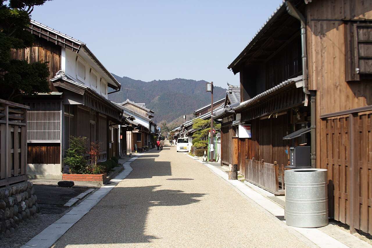 A straight paved street lined with old wooden townhouses with latticed windows and tiled eaves, leading toward forested hills in Seki-juku, Kameyama.