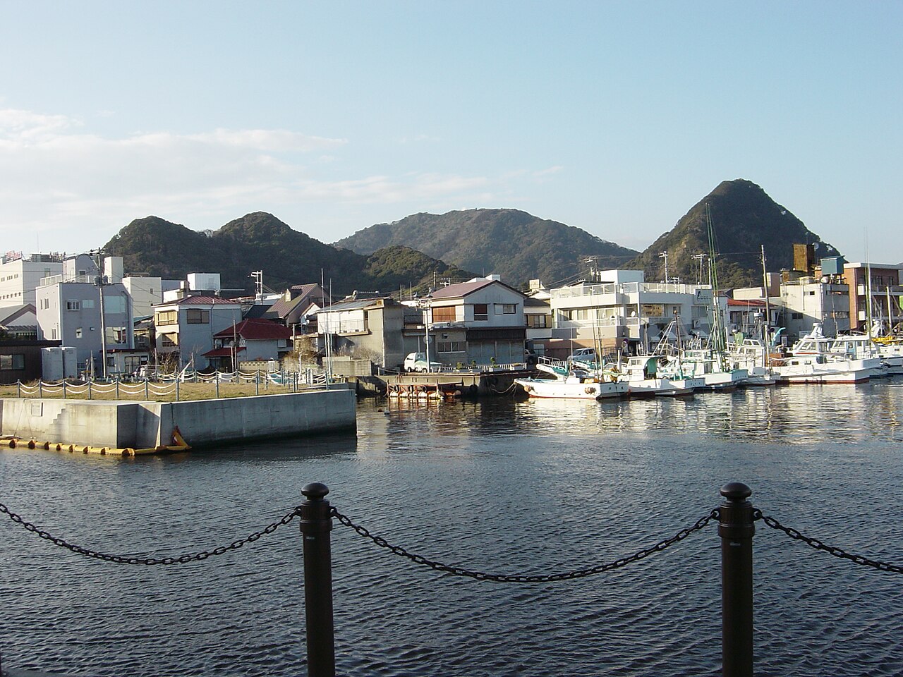 Small fishing boats moored along a harbor quay with low houses and green hills behind, seen across the water and a chain railing.