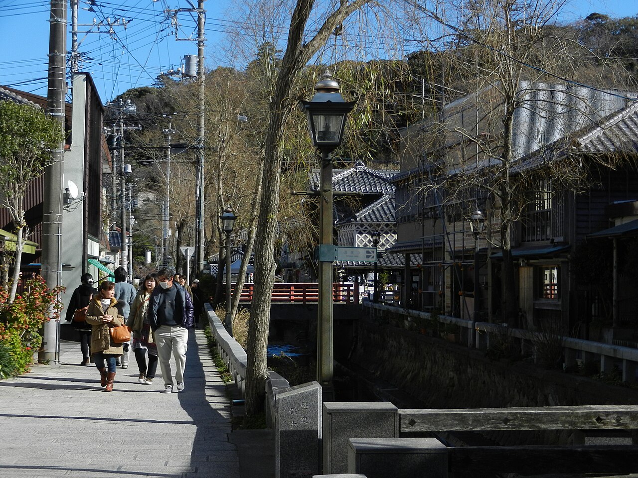 A lane beside a stone-walled canal with a street lamp, people walking, and old wooden buildings along the far side.