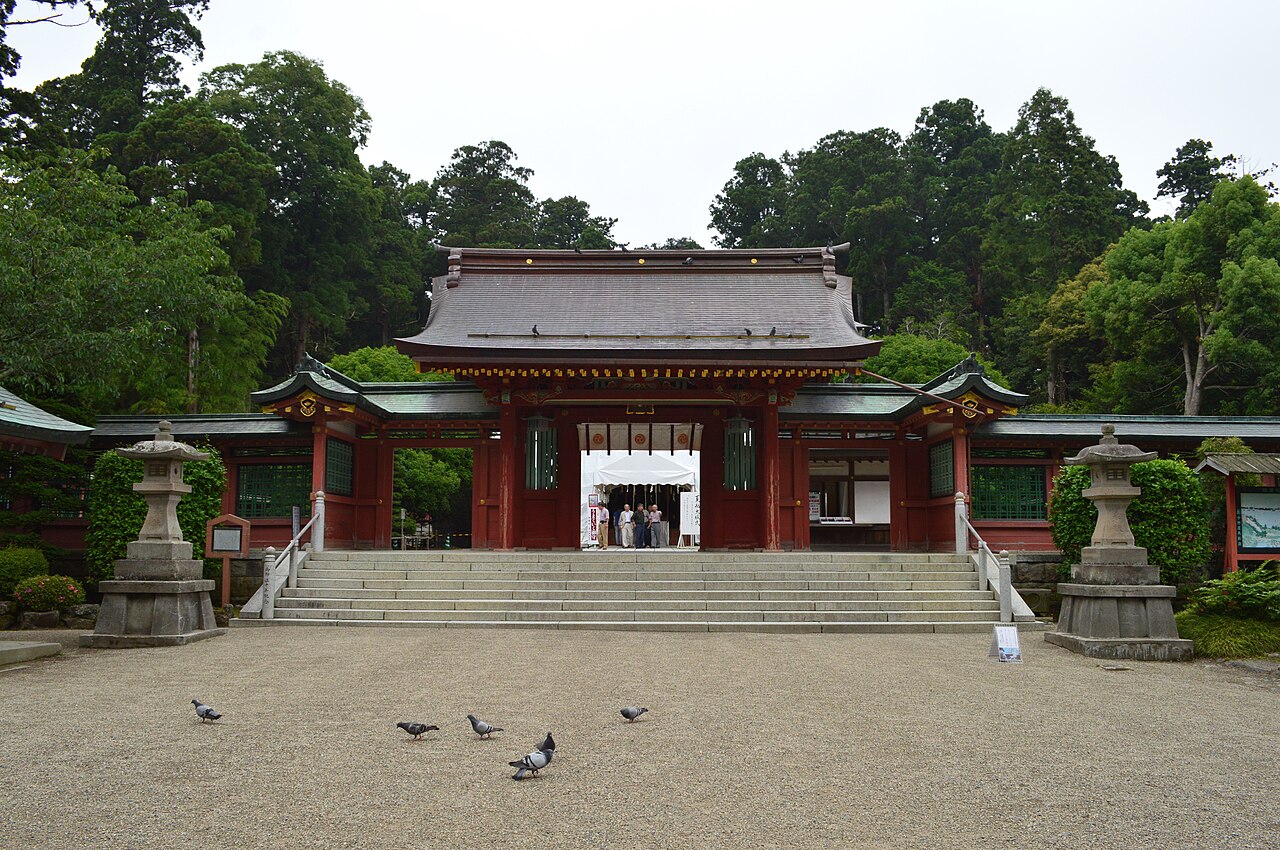 The vermilion gate of Shiwahiko Shrine behind a gravel courtyard, with stone lanterns and tall cedars on either side