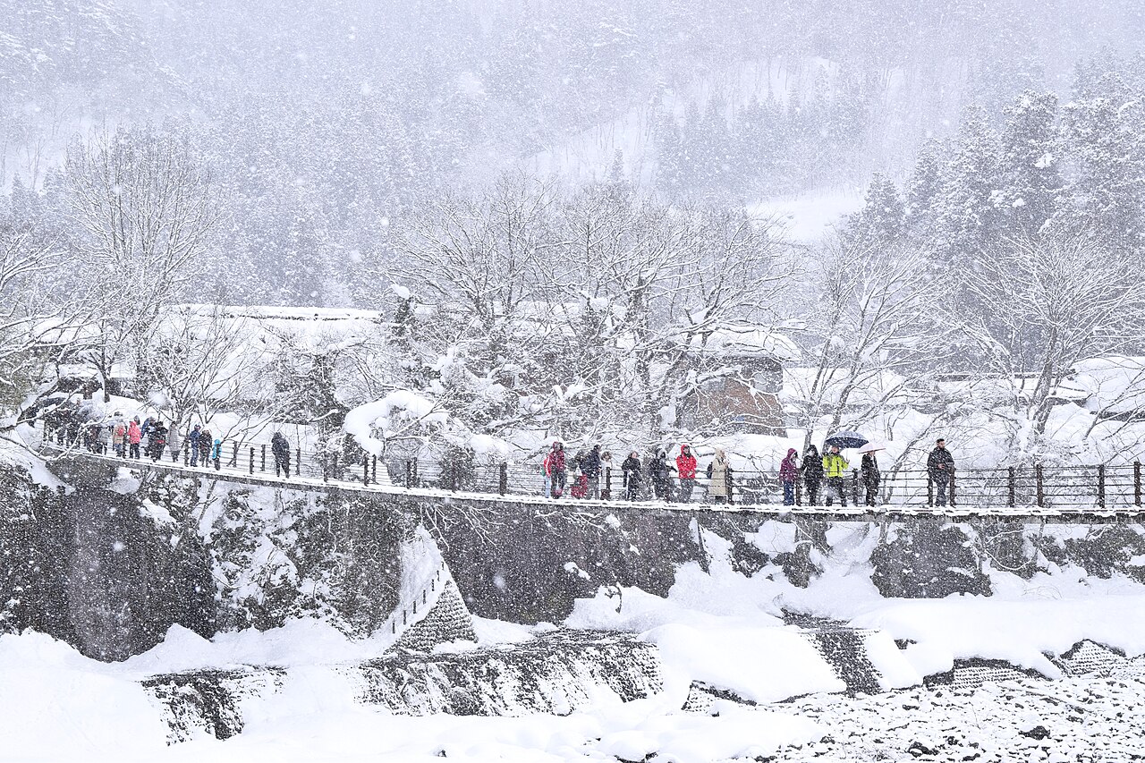 Visitors crossing a long footbridge over a snowy river gorge in heavy snowfall, with snow-laden trees and thatched roofs behind