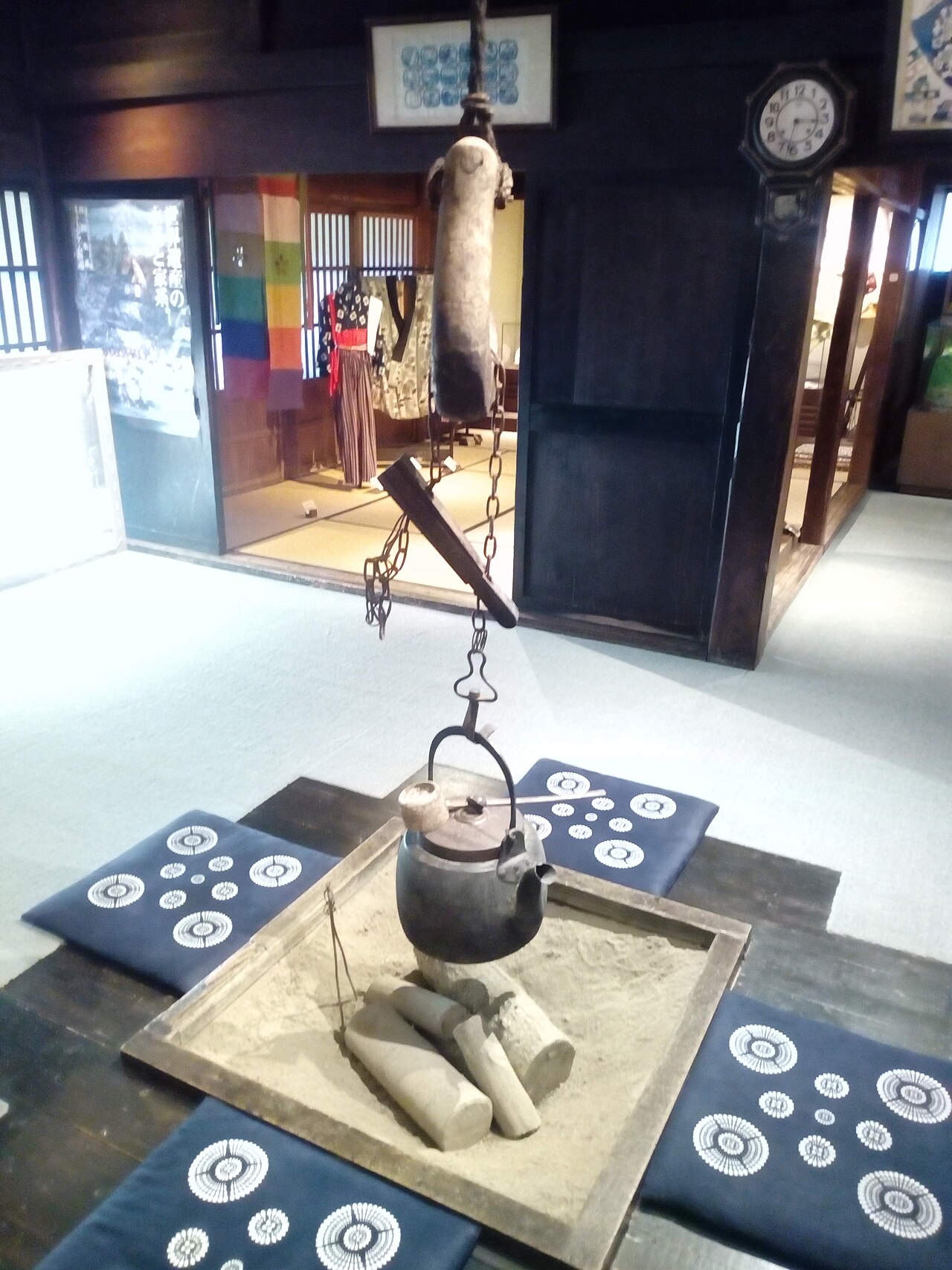 A sunken irori hearth with an iron kettle hanging from a pot hook over logs, surrounded by navy floor cushions inside a gassho-zukuri house