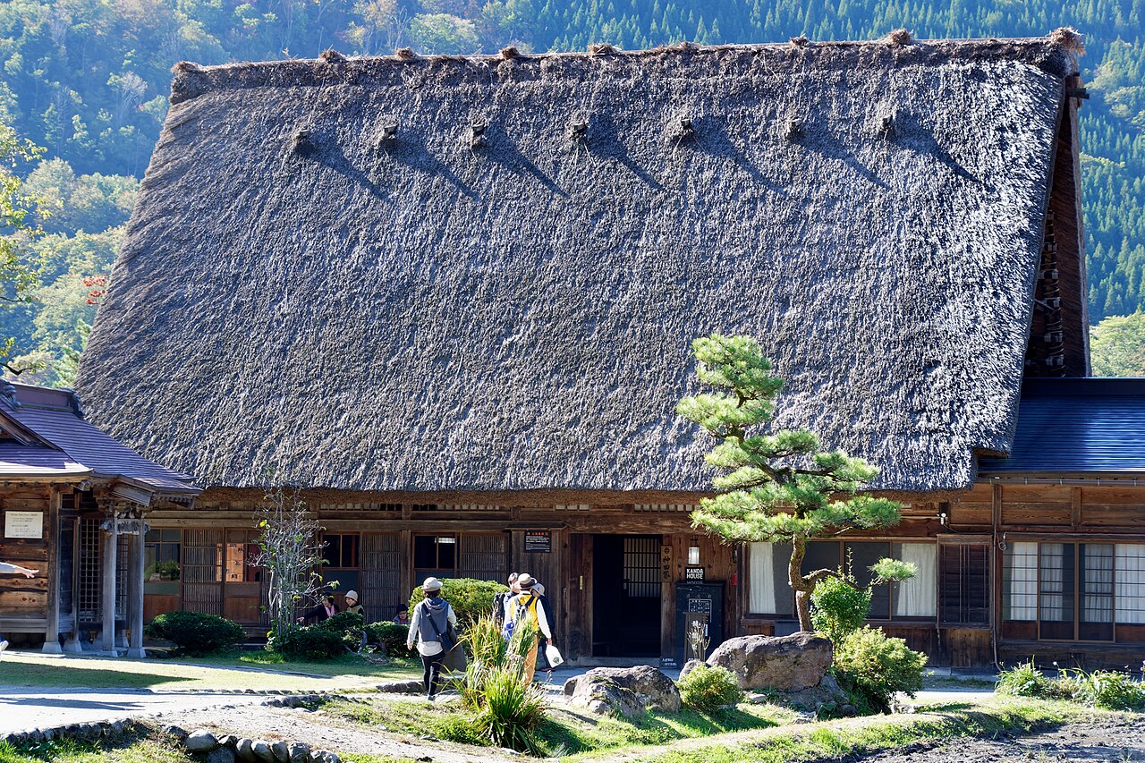 The sharply pitched thatched roof of Kanda House with visitors walking past its entrance and a pine tree in the garden, forested hills behind