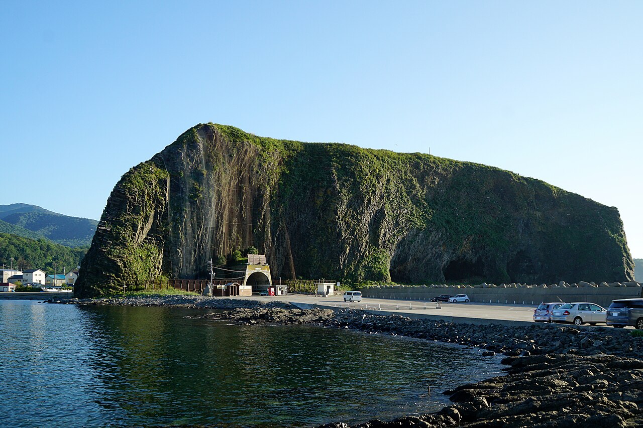 A massive dome of rock with a road tunnel at its base rises above a calm harbor bay at Utoro.