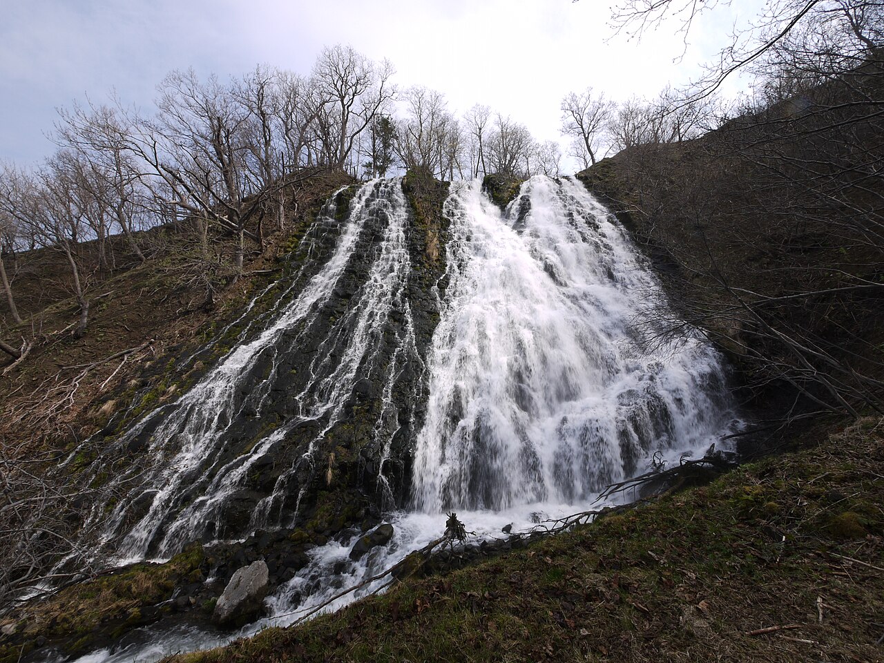 A wide fan-shaped waterfall spreads down a rocky slope among bare spring trees.