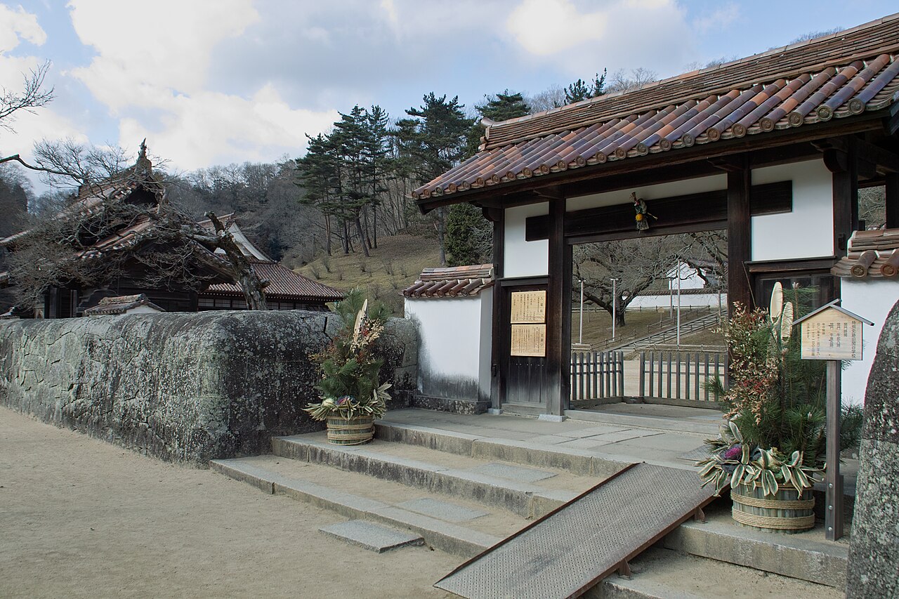 Tiled gate of Shizutani School beside a rough stone wall, with a New Year pine and bamboo decoration at the steps