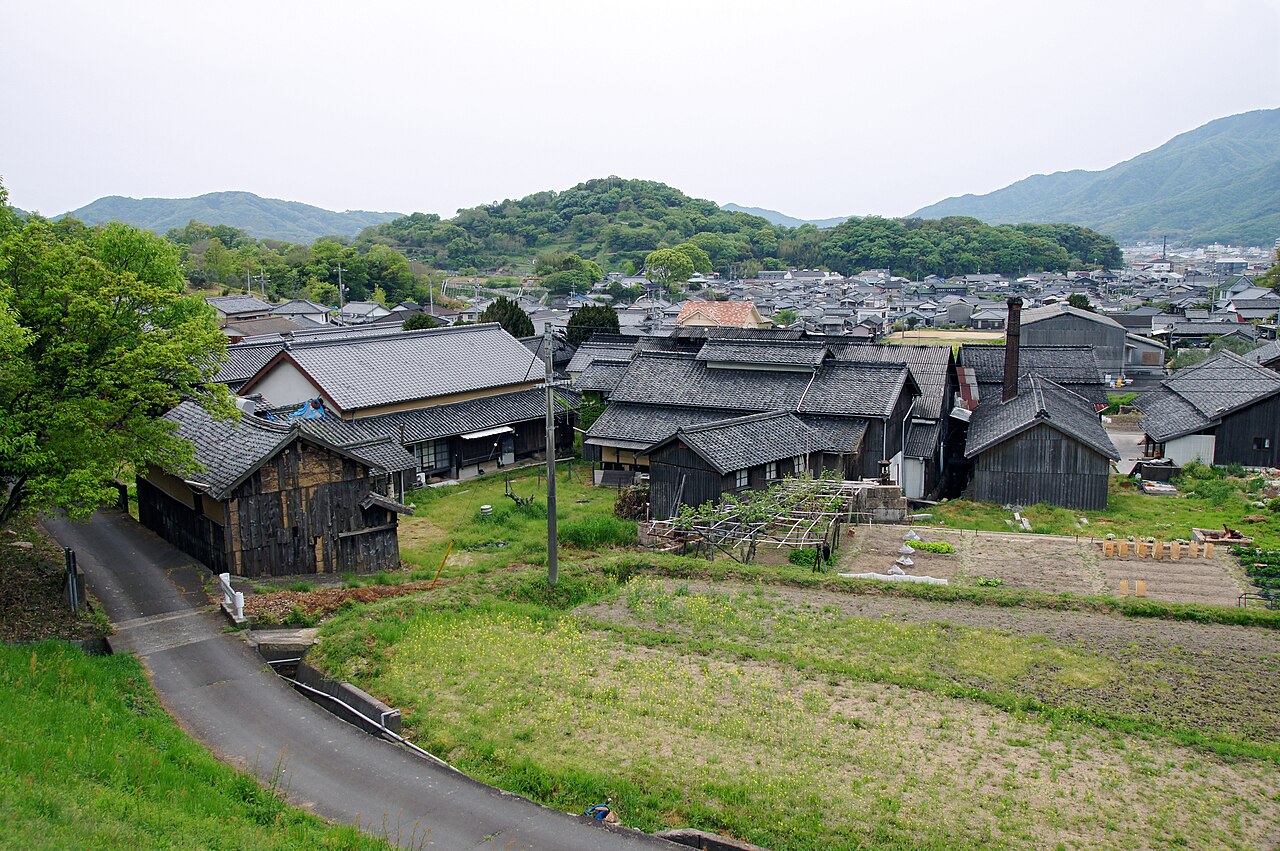 Dark-timbered storehouses and tiled roofs of the Hishio-no-sato soy sauce district, with a village and green hills beyond.