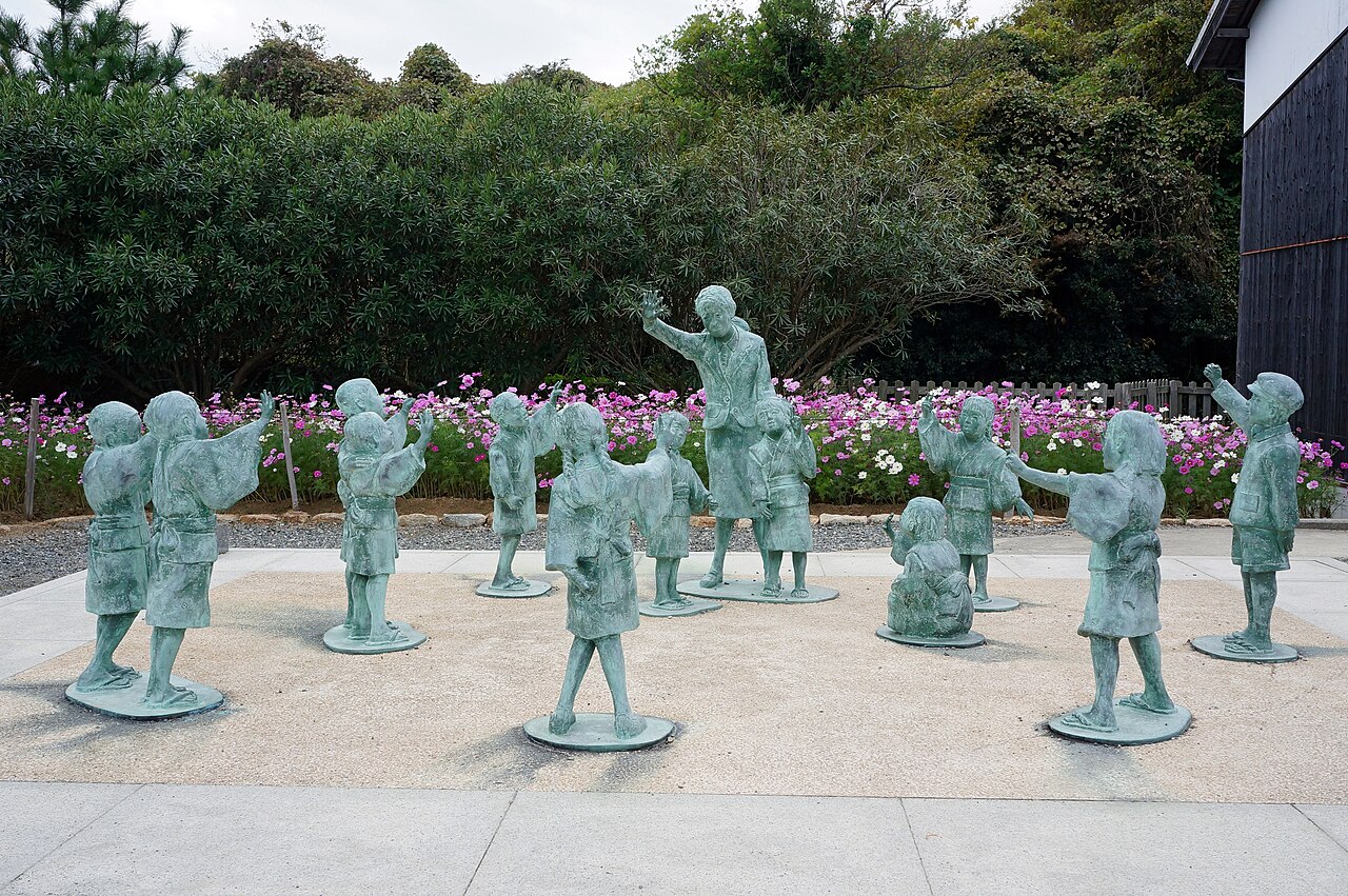 Bronze statues of a teacher and schoolchildren with raised arms, with pink cosmos flowers and dark wooden buildings behind, at the Twenty-Four Eyes Movie Village.