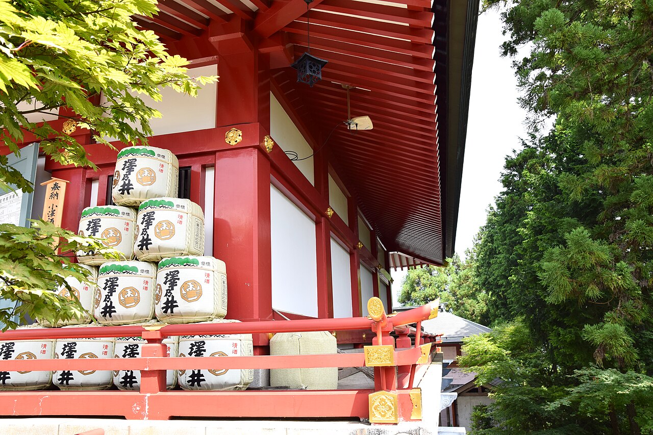 Stacked sake barrels beside the vermilion hall of Musashi Mitake Shrine, framed by maple leaves.