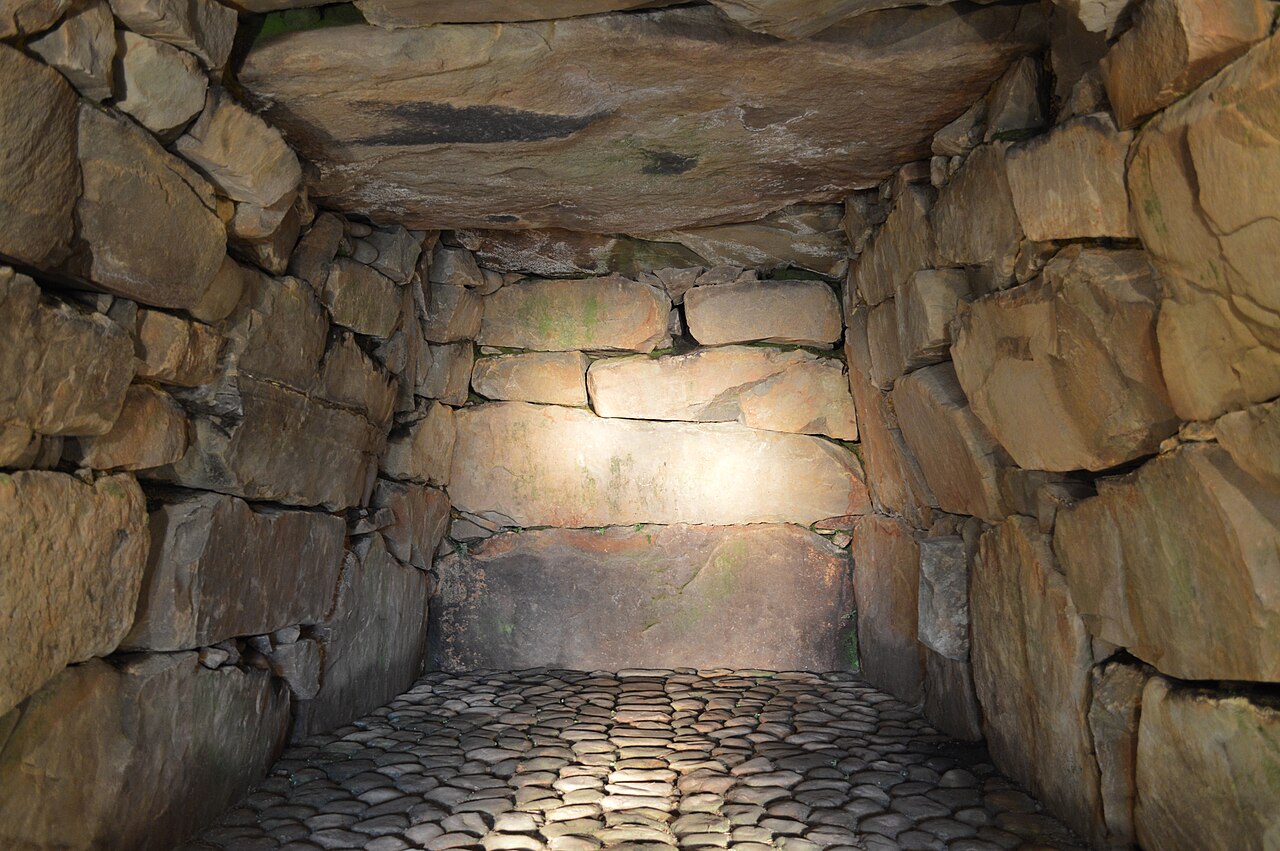 Inside a burial chamber, walls of fitted rough stones rise to a flat stone ceiling above a floor paved with rounded cobbles, with light reaching the far wall