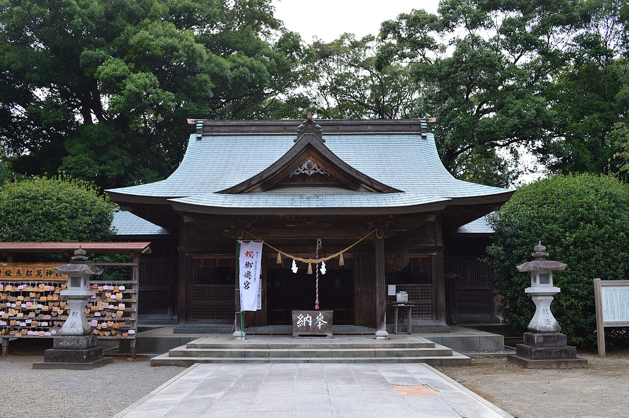 A wooden shrine worship hall with a pale grey-green roof and a rope with paper streamers hung across the front, stone lanterns at either side and wooden prayer plaques stacked at left