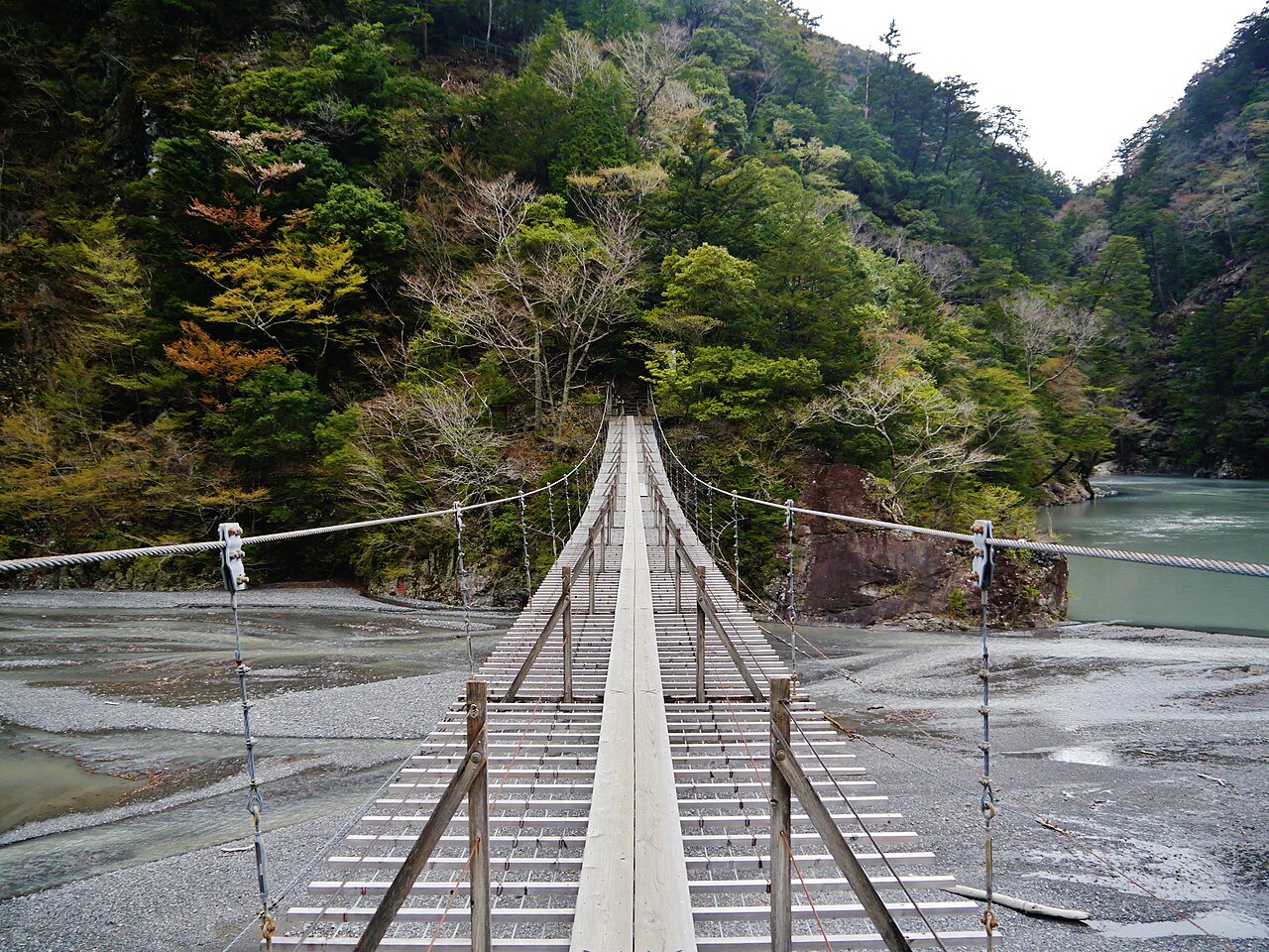 Looking straight along the plank deck of the Yume no Tsuribashi toward a wooded slope, with grey riverbed and pale green water on both sides
