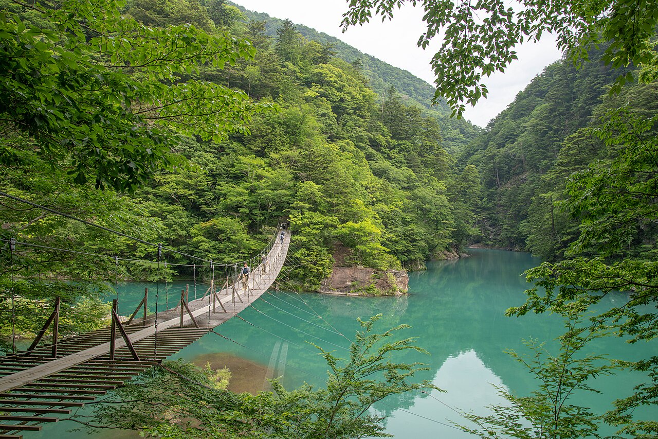 A narrow wooden suspension bridge crossing turquoise water between forested slopes in Sumatakyo Gorge
