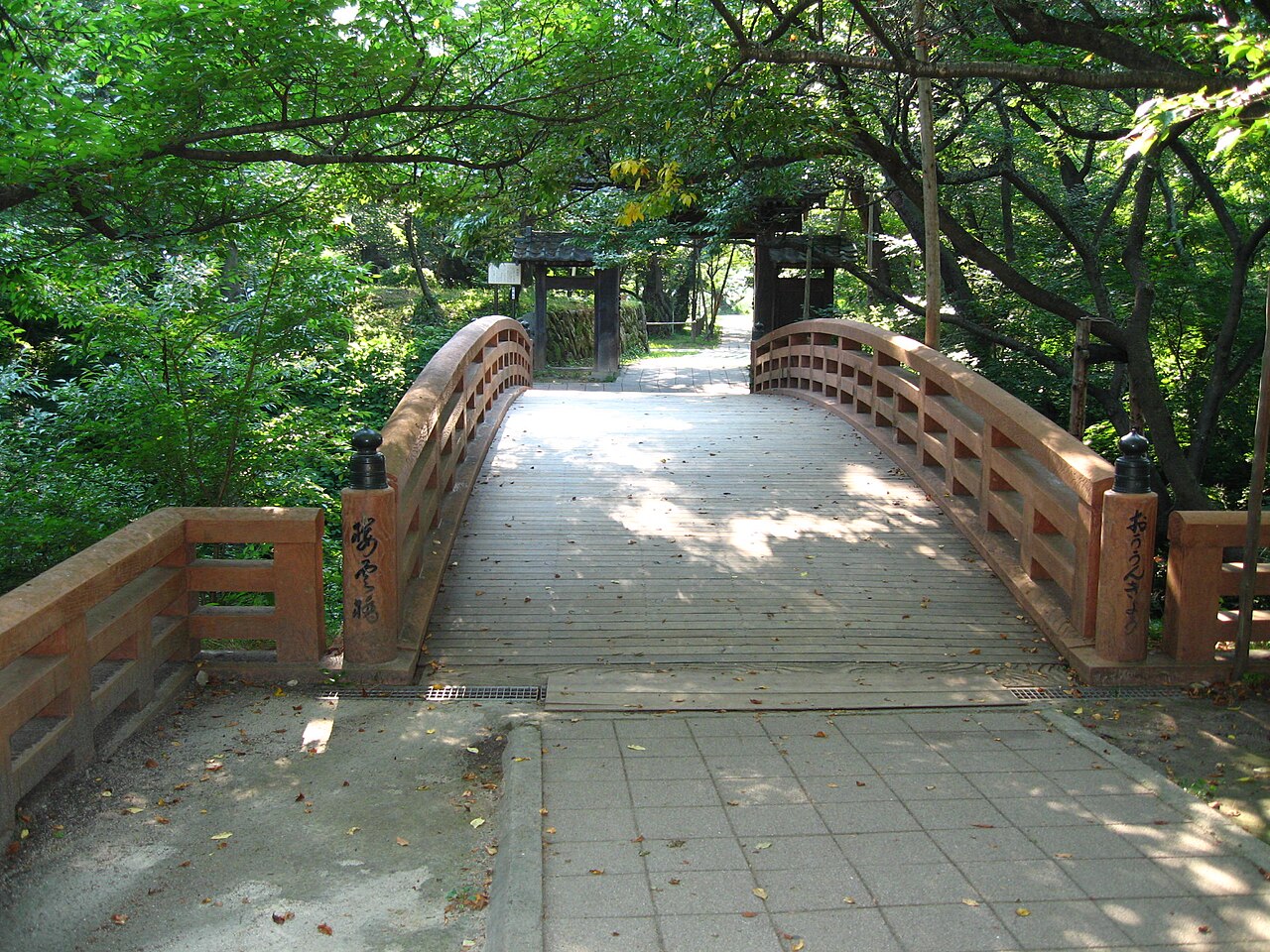 The arched vermilion wooden Ounkyo bridge inside Takato Castle Ruins Park, leading under green summer trees toward a small wooden gate.
