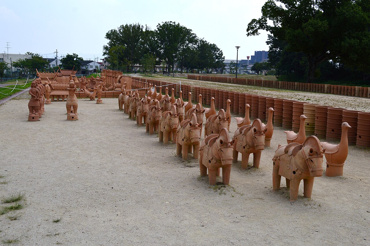Rows of reddish-brown clay horse figures in harness and long-necked bird figures standing on a sandy ground, with rows of cylindrical clay pipes behind them and tall trees and houses at the edge of the park.