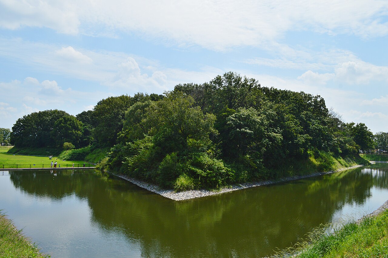 A large wooded mound rising from a still moat that mirrors the trees, with a grassy bank on the left where a few tiny visitors stand, under a blue sky with scattered clouds.