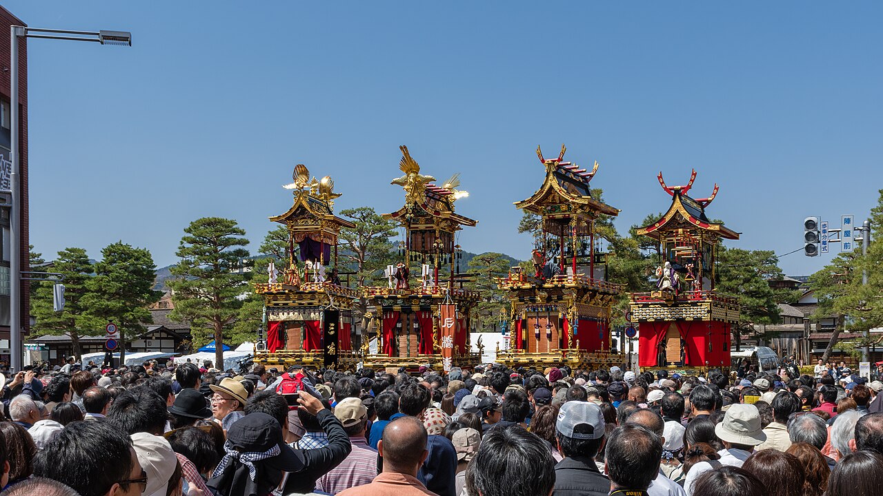 Four ornate festival floats (yatai) with gold carvings and red curtains lined up in front of a dense crowd during the Takayama Matsuri.